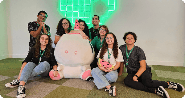 Grupo de jovens sorrindo ao redor de um mascote de pelúcia em ambiente interno.